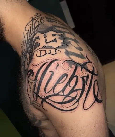 Geschwungenes Chicano-Lettering „Liebste“ über die Schulter tätowiert
